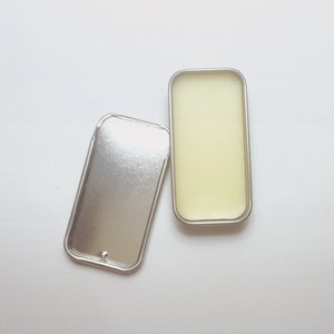 Op de afbeelding: Een zilveren blik met een deksel, open om een ​​gele balsem erin te laten zien.