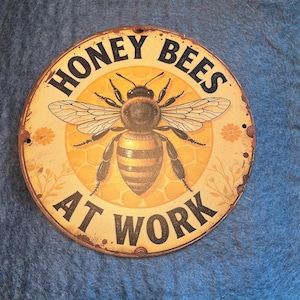 May include: Round metal sign with a bee illustration and the text "HONEY BEES AT WORK." The sign has a yellow and brown color scheme with a distressed, vintage look. The bee is centered with detailed wings.