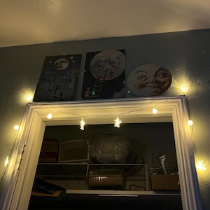 May include: Three celestial-themed wall art pieces above a doorway. One piece has the text "Sometimes I Just Look Up...". A string of star-shaped lights and round lights are strung above the doorway. The walls are painted a muted teal color.