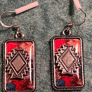 May include: A pair of rectangular earrings with a silver-colored frame and a red and patterned background. Each earring features a silver-colored diamond shape in the center, surrounded by a zig-zag design. The earrings have silver-colored hooks.