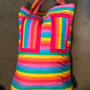May include: A quilted tote bag with a vibrant rainbow striped pattern. The bag features two rectangular pockets outlined in bright pink. The handles and the top edge of the bag are also in a rainbow color scheme.