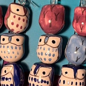 May include: Close-up of several ceramic owl earrings in various colors. The owls are painted in shades of blue, white, and burgundy, with black eyes and decorative patterns. The earrings are displayed against a turquoise background.