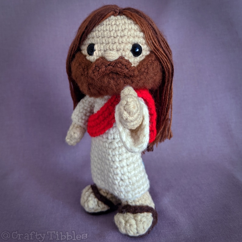 May include: Crocheted amigurumi doll of Jesus with brown hair, a white robe, and a red sash. The doll has a brown beard and mustache and is holding out its right hand.