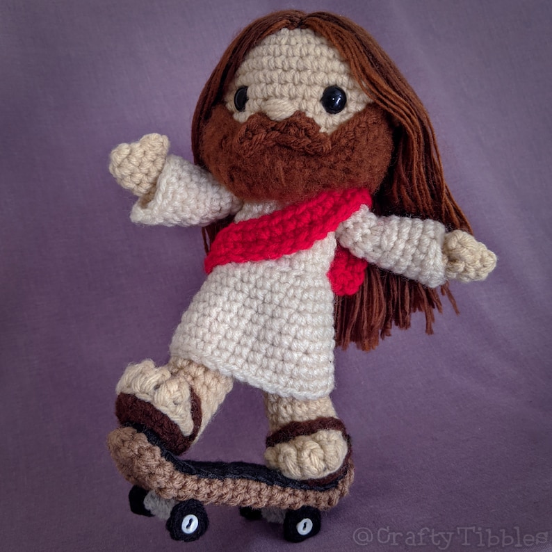 May include: A crocheted figure of Jesus wearing a red robe and brown hair, riding a miniature skateboard.