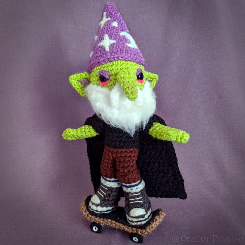 May include: A green crocheted gnome wearing a purple hat with white stars and a black cape is riding a miniature skateboard with black wheels. The gnome has a white beard and red eyes.