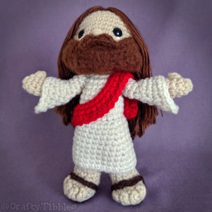 May include: Crocheted amigurumi doll of a person wearing a white robe and a red sash. The doll has brown hair and a brown beard.