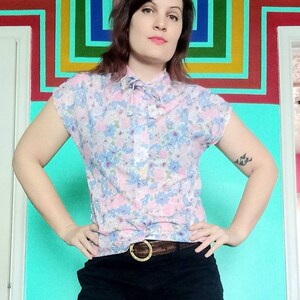 Può includere: Una donna indossa una blusa rosa con stampa floreale, un fiocco, pantaloncini neri e una cintura dorata. È in piedi davanti a un muro con un motivo arcobaleno.