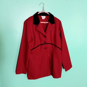 May include: A red blazer with a black velvet collar and black trim. The blazer has three black buttons down the front and is hanging on a patterned hanger against a teal wall.