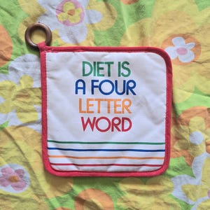 Puede incluir: Salvamanteles cuadrado blanco con el texto "DIET IS A FOUR LETTER WORD" en verde, azul, naranja y rojo. El salvamanteles tiene un borde rojo, rayas arcoíris y un anillo de madera para colgar.