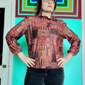 Puede incluir: Una mujer con una blusa de estampado rojo, marrón y azul con cuello de volantes y pantalones vaqueros negros. Está de pie frente a una pared turquesa con rayas de arcoíris.