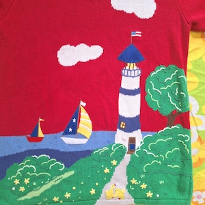 May include: Red knitted sweater with a lighthouse, two sailboats, clouds, and green bushes embroidered on it. The lighthouse has a blue roof and a small American flag on top. There are yellow flowers embroidered on the ground.