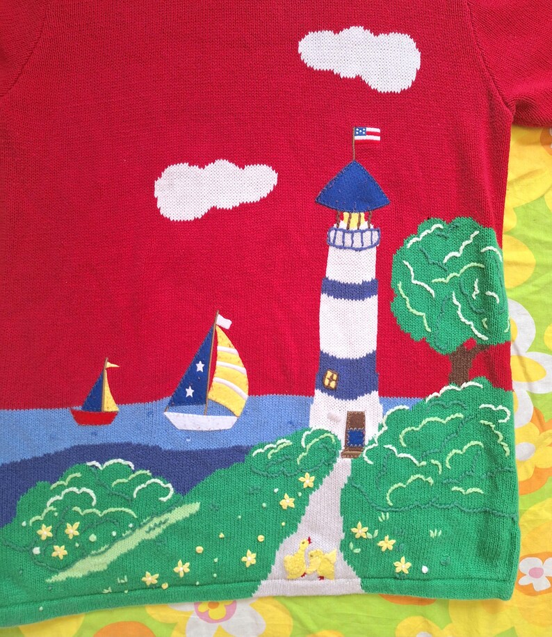 May include: Red knitted sweater with a lighthouse, two sailboats, clouds, and green bushes embroidered on it. The lighthouse has a blue roof and a small American flag on top. There are yellow flowers embroidered on the ground.