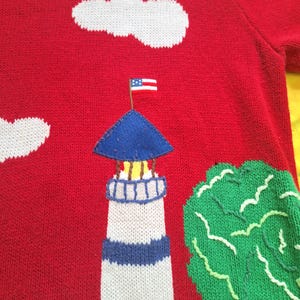 May include: Red knitted sweater with a white lighthouse, blue roof, and an American flag on top. There is a green tree and white clouds in the background.
