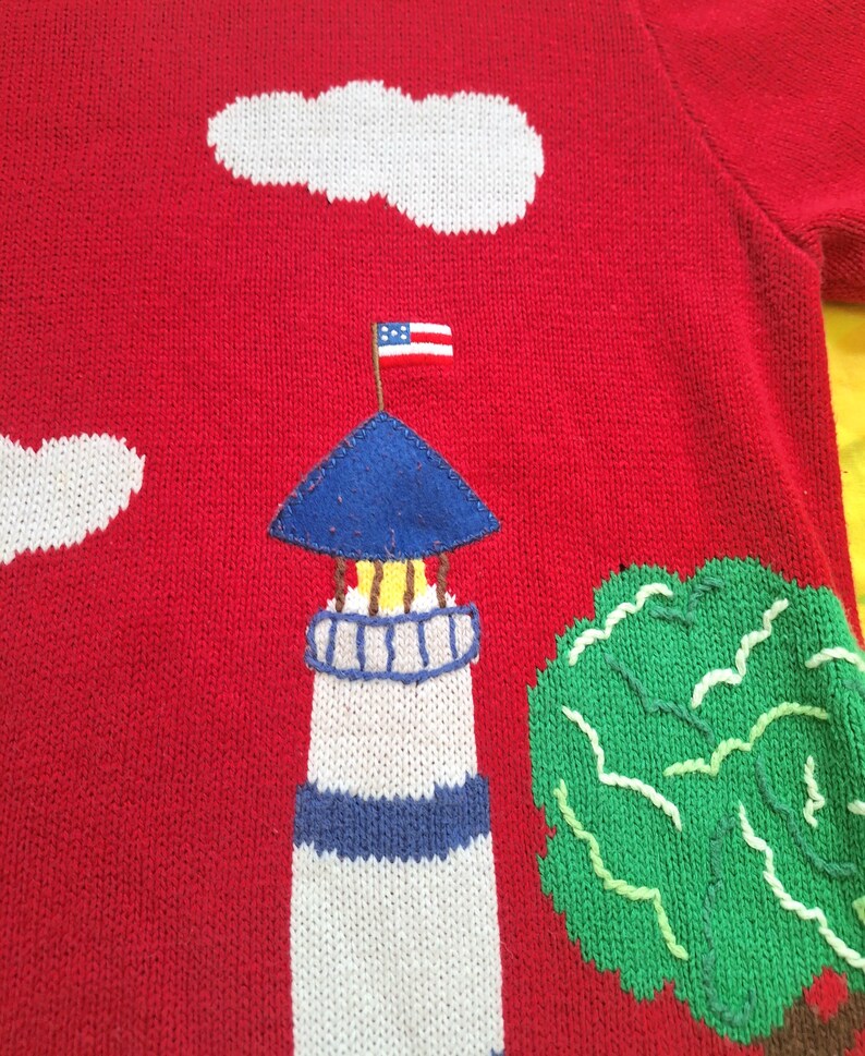 May include: Red knitted sweater with a white lighthouse, blue roof, and an American flag on top. There is a green tree and white clouds in the background.