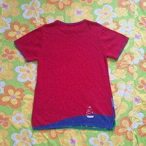 May include: Red short-sleeved knit shirt with a blue and green trim and a small sailboat design on the bottom.