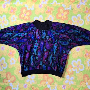 May include: A vintage sweater with a black collar and cuffs. The sweater features a vibrant paisley pattern in shades of purple, blue, and pink on a black background. The sleeves are wide and the overall style is loose and comfortable.