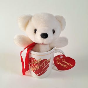 Könnte beinhalten: Weißer Plüschteddybär, der in einer weißen Keramiktasse mit einem roten Herzen und goldenen Schriftzügen sitzt, auf denen "Happy Valentine's Day" steht. Ein rotes Band ist um den Hals des Bären gebunden. Ein rotes herzförmiges Etikett mit goldenen Schriftzügen sagt "Für jemanden Besonderes!"