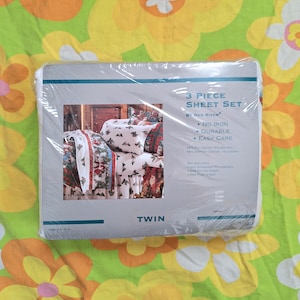 May include: A white package with a picture of a twin-sized bed with a Christmas-themed sheet set. The package says "3 Piece Sheet Set by Dan River" and lists features like "No Iron", "Durable", and "Easy Care".