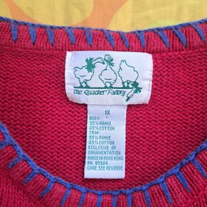 May include: Red sweater with blue trim and a white tag with green text that reads "the Quacker Factory" and "1X". The tag also includes the fabric content: "Body: 55% Ramie 45% Cotton Trim: 55% Ramie 45% Cotton Exclusive of Ornamentation Made in Hong Kong RN 95524 Care See Reverse"