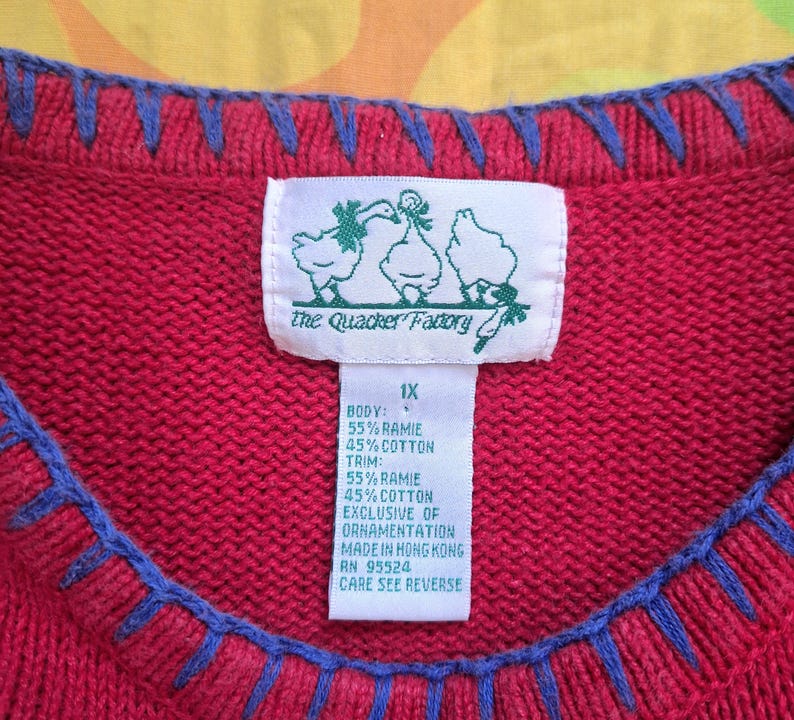 May include: Red sweater with blue trim and a white tag with green text that reads "the Quacker Factory" and "1X". The tag also includes the fabric content: "Body: 55% Ramie 45% Cotton Trim: 55% Ramie 45% Cotton Exclusive of Ornamentation Made in Hong Kong RN 95524 Care See Reverse"