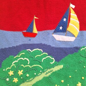 May include: A red, blue, and yellow knitted sweater with a design of two sailboats and a green island with yellow flowers.