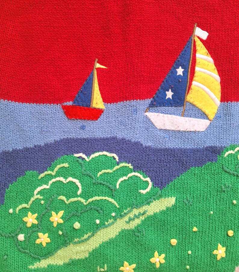 May include: A red, blue, and yellow knitted sweater with a design of two sailboats and a green island with yellow flowers.