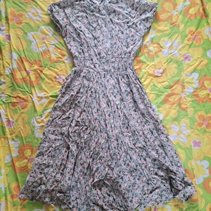 May include: A vintage floral print dress with short sleeves and a gathered waist. The dress features a pattern of small pink and green flowers on a light background. The skirt is pleated, and the dress is made of a lightweight fabric.