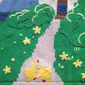 May include: A knitted fabric with a green background and a white path. The path has two yellow embroidered chicks facing each other. There are yellow embroidered flowers throughout the fabric.