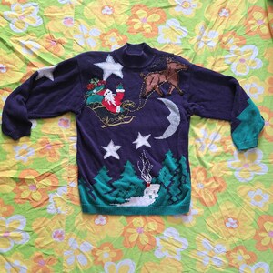 Può includere: Un maglione di Natale blu a maglia con un Babbo Natale verde, rosso e bianco che cavalca una slitta trainata da una renna. Il maglione ha un design a stella e mezzaluna.