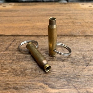 May include: Two brass bullet casing keychains on a wooden surface. One keychain is lying down, and the other is standing upright. The keychains have silver key rings attached. The background is a wooden surface.