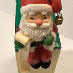 May include: A ceramic figurine of Santa Claus holding a scroll and a bell. He is wearing a red hat, white beard, and red and white suit. The figurine is sitting on a green and white box.