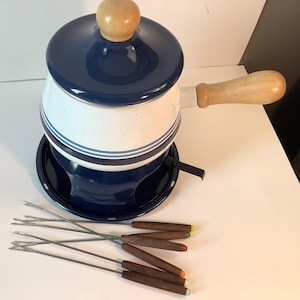 May include: Blue and white enamel fondue pot with a wooden handle and lid. The pot sits on a blue metal base with a removable warming element. Six fondue forks with wooden handles are included.