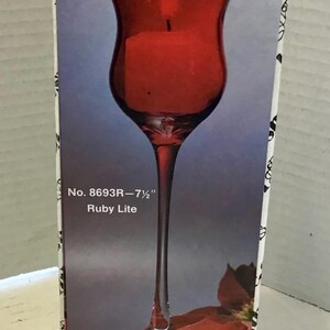 May include: A tall, ruby red glass candle holder with a slender stem, 7.5 inches tall, on a box. The box has the text "No. 8693R-7 1/2" Ruby Lite". A red flower is visible at the bottom.