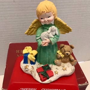 May include: A ceramic Christmas angel figurine with blonde hair and yellow wings, holding a white dog. The angel is wearing a green dress and is surrounded by a blue gift with a yellow bow, a brown teddy bear, and a red gift box.