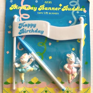 May include: A vintage Avon "Birthday Banner Buddies" set. The set includes a white banner with "Happy Birthday" in blue script, two ballerina bunny figurines, and a blue and white pole. The packaging is blue with yellow and pink accents.