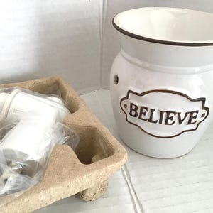 May include: A white ceramic oil warmer with a brown rim and the word "BELIEVE" in brown lettering. The warmer has a round shape with a wide opening and two small holes. A white plug-in warmer is in a cardboard tray.