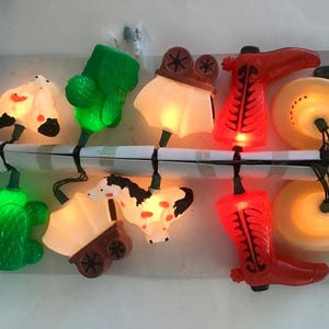 May include: String of novelty lights featuring cowboy-themed shapes. The string includes lights shaped like cacti, a horse-drawn wagon, a horse, a film reel, cowboy boots, and cowboy hats. The lights are in various colors, including green, red, and yellow.