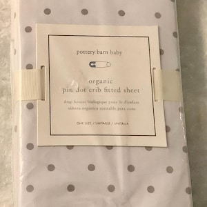 May include: A white organic cotton crib fitted sheet with a gray pin dot pattern. The sheet is packaged in plastic and has a label that reads "pottery barn baby organic pin dot crib fitted sheet".