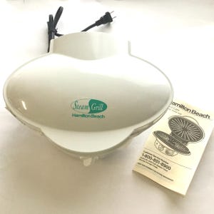 May include: A white Hamilton Beach Steam Grill with a black power cord. The grill has the words "Steam Grill" and "Hamilton Beach" printed on the front. A product manual is also included.