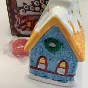 May include: A ceramic Christmas house candle holder with a blue stone-like facade, orange roof trim, and a white snowy roof. The house has two arched windows and a green wreath. A red candle is in the foreground, and a box with a similar house design is in the background.