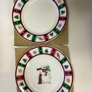 May include: Two white ceramic plates with a festive winter design. Each plate features a border with alternating green trees, red stripes, and winter-themed icons. One plate has a polar bear graphic.