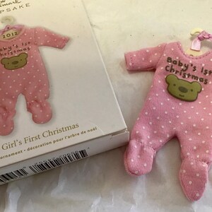 May include: Pink baby's first Christmas ornament shaped like a onesie, with white polka dots and a green bear applique. The ornament has the text "Baby's 1st Christmas" and the year "2012".