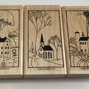 May include: Three wooden stamps with black ink illustrations of rural scenes. The stamps depict a house, church, and windmill, with trees, flowers, and a snowman. The text "© 2003 STAMPIN' UP!" is printed on each stamp.