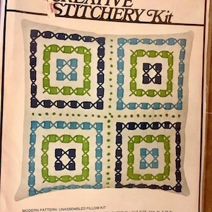 May include: A vintage crewel embroidery kit for a pillow featuring a geometric design with blue, green, and white colors. The kit includes the fabric, yarn, needle, and instructions. The finished pillow size is 13 inches by 13 inches.
