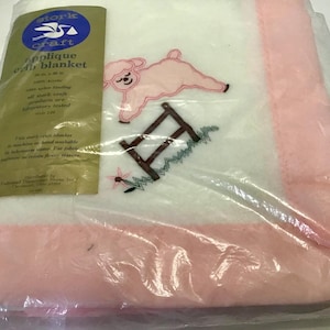 May include: A new baby blanket in a plastic package. The white blanket has a pink border and an embroidered design of a pink lamb, a brown fence, and a flower. The packaging includes the text "Stork Craft Applique Baby Blanket".