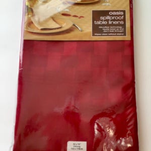 May include: A red spillproof table linen, packaged in plastic. The label reads "oasis spillproof table linens". The dimensions are 60 x 78 inches (152 x 198 cm). The fabric is a deep red with a subtle checkered pattern.