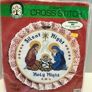May include: A stamped cross stitch kit featuring a Christmas scene with the text "Silent Night Holy Night." The kit includes a cross-stitch design with a circular frame, a white lace trim, and a red background.