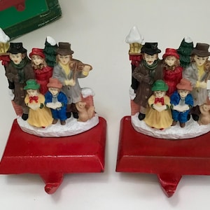 May include: Two Christmas stocking holders with a festive scene. Each red holder features a group of carolers, including a violinist, children, and a small dog, all set against a snowy backdrop. The figures are dressed in winter attire.