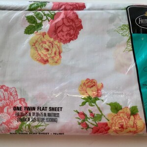 May include: A white twin flat sheet in a plastic package, featuring a floral pattern of large pink and yellow roses with green leaves. The package has text that reads "Fruit of the Loom" and "no iron Muslin".