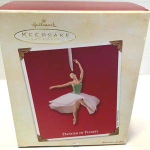 May include: Hallmark Keepsake ornament featuring a ballerina in a green and white tutu, arms outstretched. The ornament is set against a red background with the text "Dancer in Flight". The box is cream-colored with gold accents.